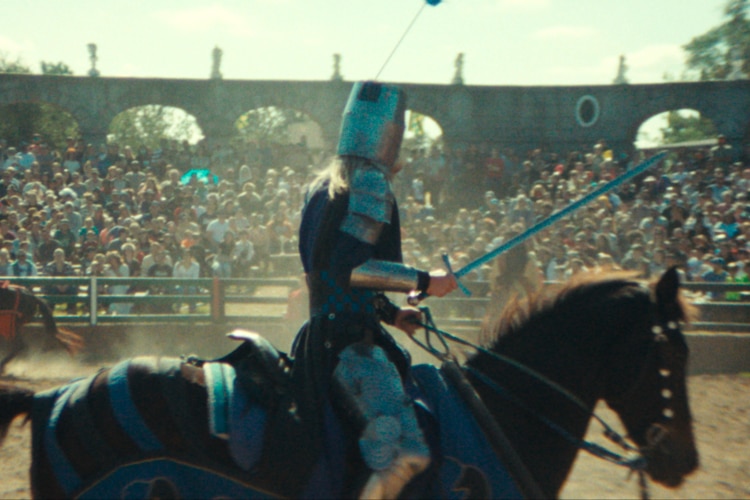 A man in armour with a sword sits on a horse, galloping around a field with crowds watching. 