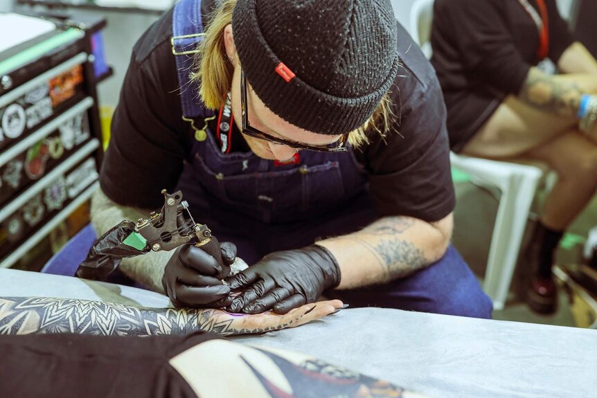 A man in a beanie tattoos a woman's hand at a bench