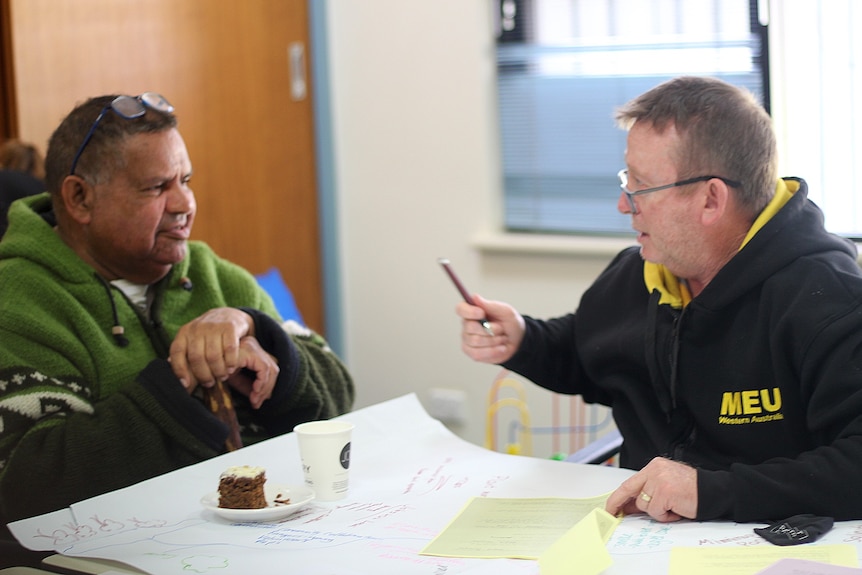 An older indigenous man and middle aged white man with a MEU shirt sit at a table talking