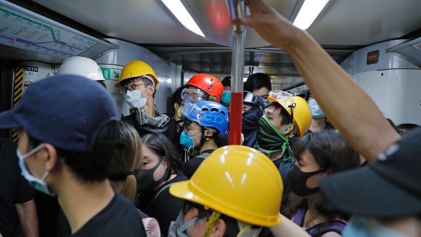 A crowded train shows a crowd of protestors wearing gas masks and coloured helmets holding metal balance poles.