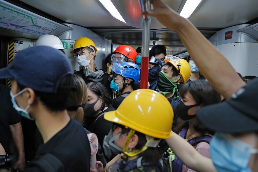 A crowded train shows a crowd of protestors wearing gas masks and coloured helmets holding metal balance poles.