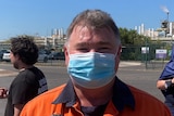 A man wearing a surgical mask and orange high-vis outside an oil refinery.