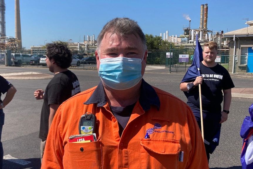 A man wearing a surgical mask and orange high-vis outside an oil refinery.