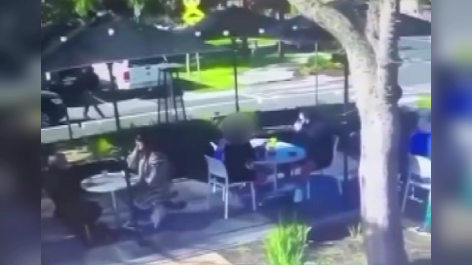 A man dressed all in black can be seen at the top left of the corner walking across a street towards people seated at a cafe.
