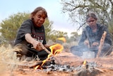 The two women sit by a campfire, one prods it with a stick