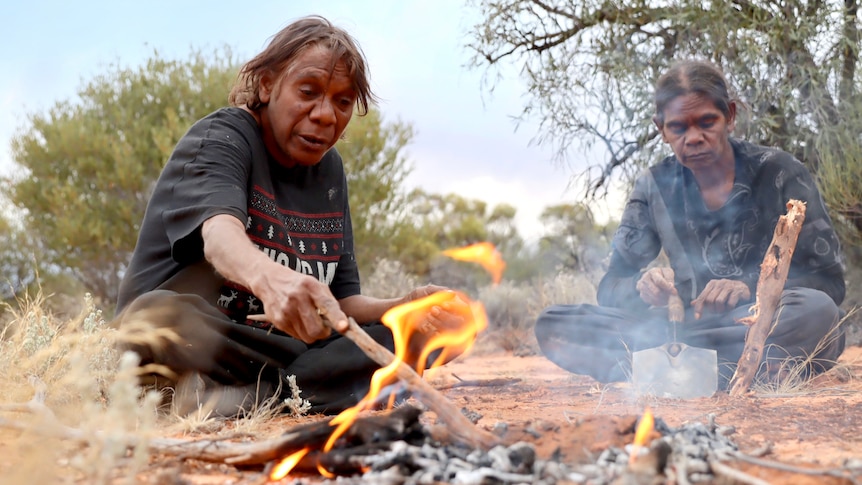 The two women sit by a campfire, one prods it with a stick