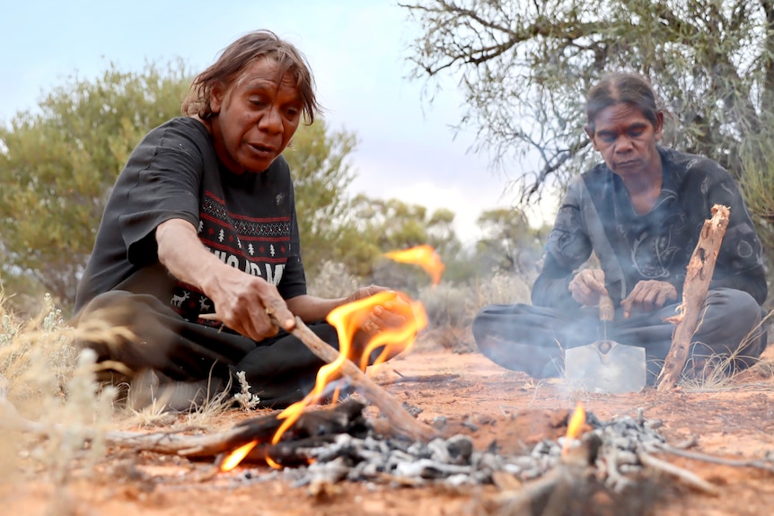 The two women sit by a campfire, one prods it with a stick