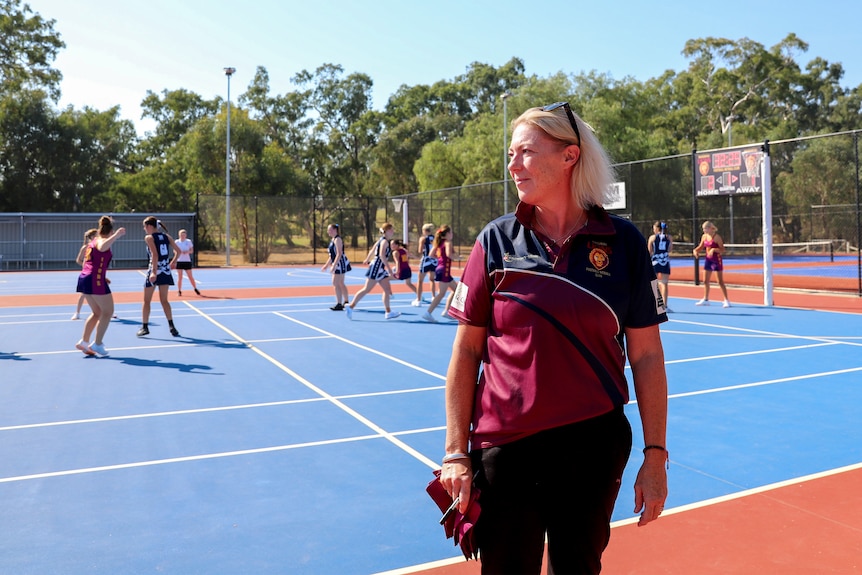 Woman wearing maroon and blue shirt stands on netball court with a women's game taking place behind her