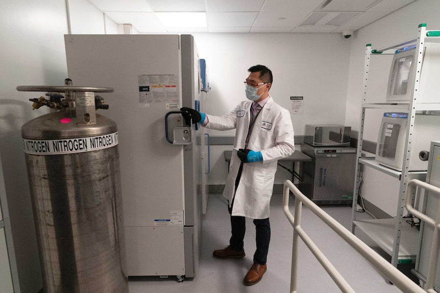 A man in a labcoat, mask and gloves stands next to a fridge.