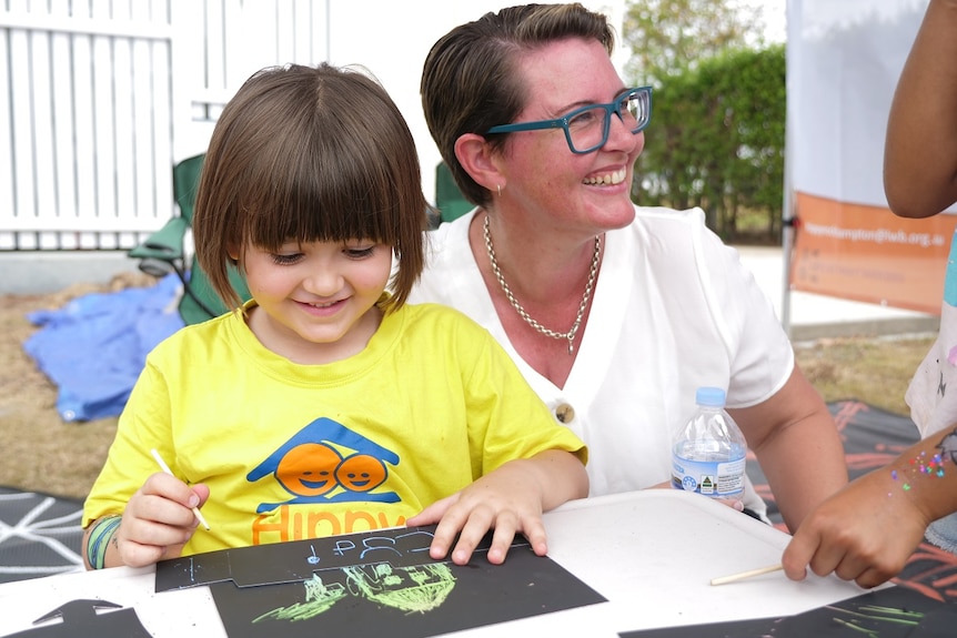 A girl with a brown bob cut and a bright yellow shirt draws with her mum, who has short brown hair and glasses.