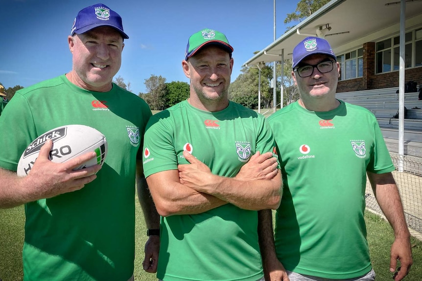 Three men stand in a row together, wearing green shirts and smiling. One holds a football. Another has his arms crossed.