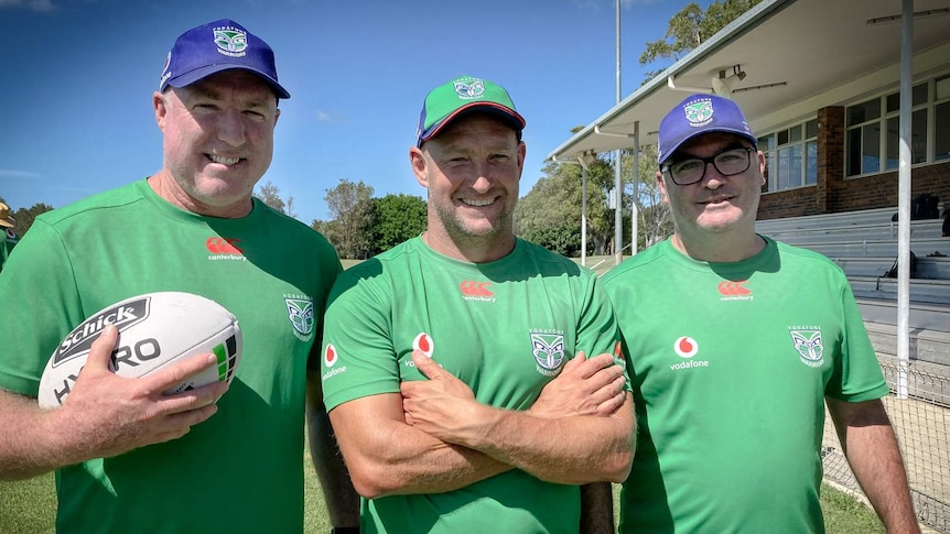 Three men stand in a row together, wearing green shirts and smiling. One holds a football. Another has his arms crossed.