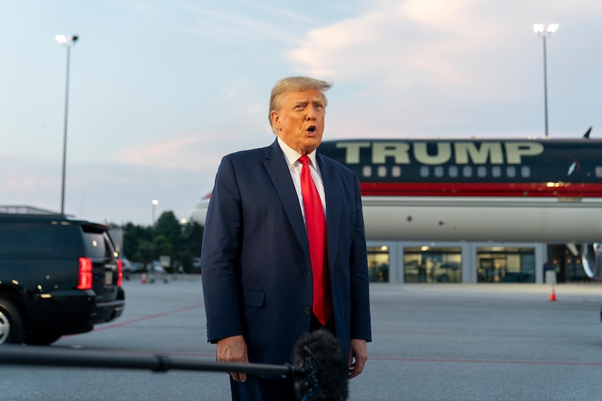 Donald Trump, in completo, parla con i giornalisti davanti a un aereo che porta il suo nome.