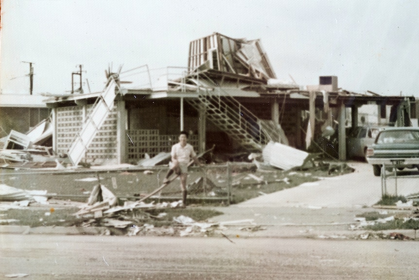 An old photograph of a man standing in front of a destroyed house after a cyclone.