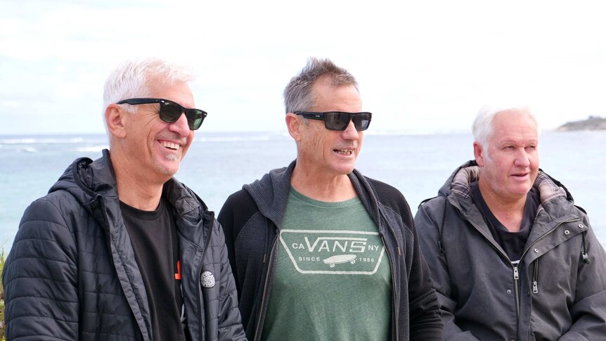 Three middle aged men stand on a beach with the ocean behind them.