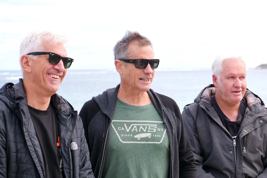Three middle aged men stand on a beach with the ocean behind them.