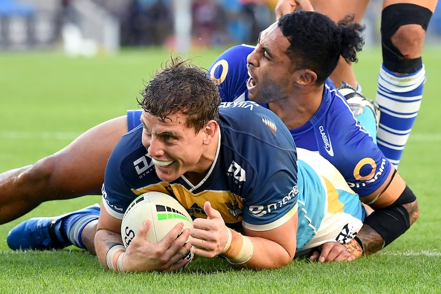 A Gold Coast Titans NRL player smiles as he holds the ball on the ground after scoring a try.