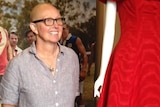 A middle-aged woman with no hair and wearing glasses stands beside a mannequin wearing a red dress from the film The Dressmaker.