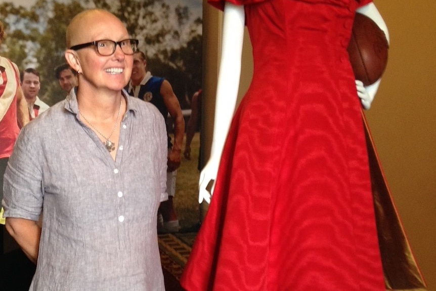 A middle-aged woman with no hair and wearing glasses stands beside a mannequin wearing a red dress from the film The Dressmaker.