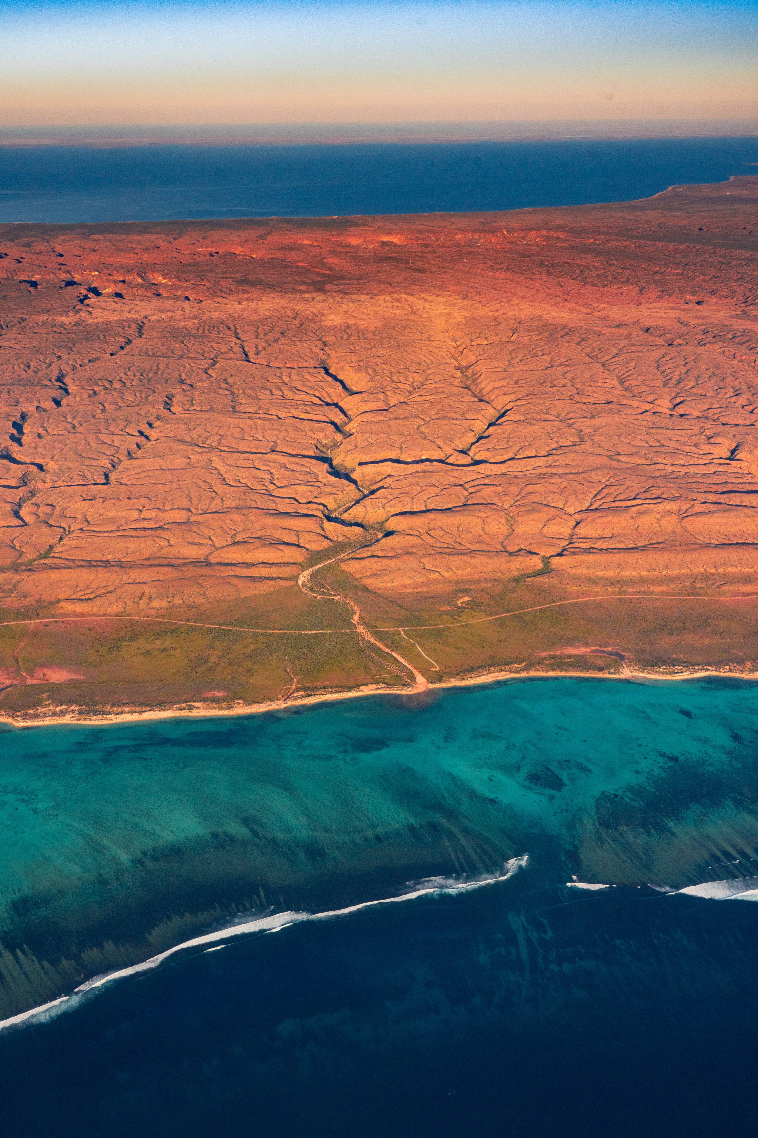 Aerial view of dark blue and turquoise waters with a reef below surface and red desert plains at dusk.