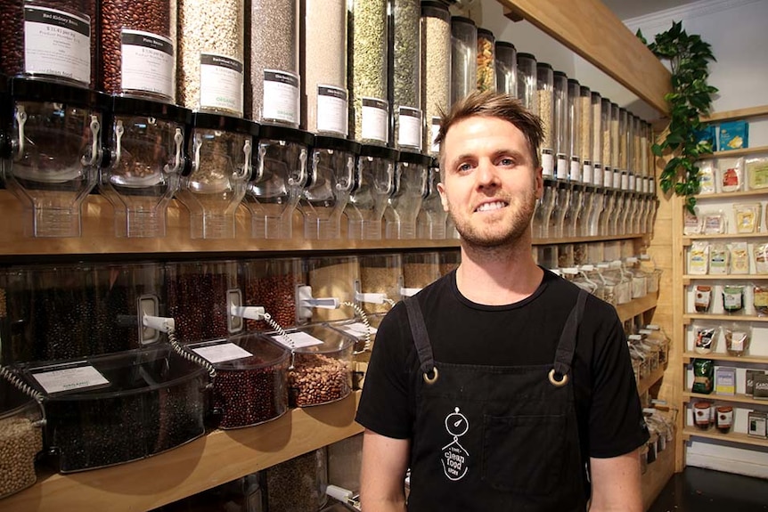 A man wearing an apron stands in a store selling whole foods, nuts and beans etc.