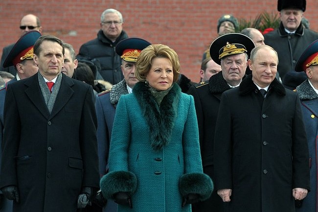 A woman in a teal coat with plush fur trims on the sleeves and neckline stands next to Vladimir Putin 