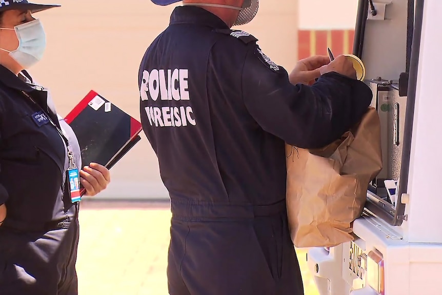 A close-up shot of police forensic officers with a brown paper bag standing at the rear of a vehicle.