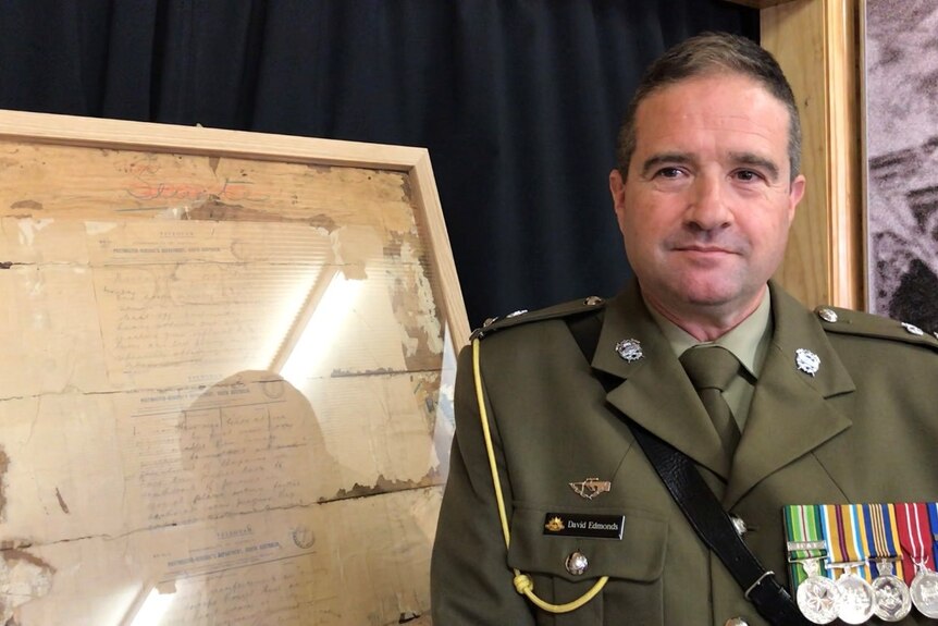 A middle-aged white male in green army uniform wearing five badges stands beside framed telegram
