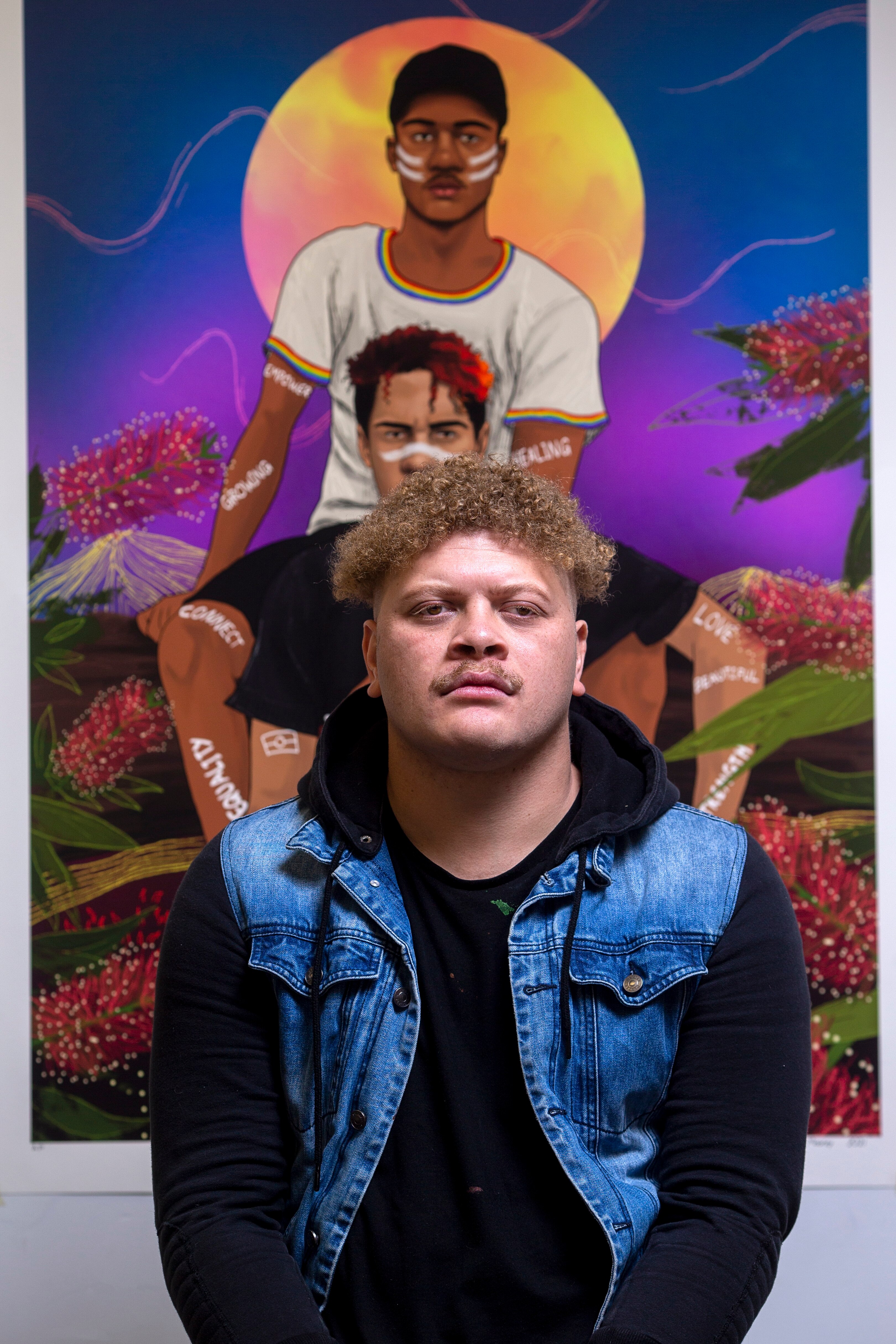 Young Indigenous Australian man with curly sandy hair and a moustache stands in front of a vividly coloured painting.