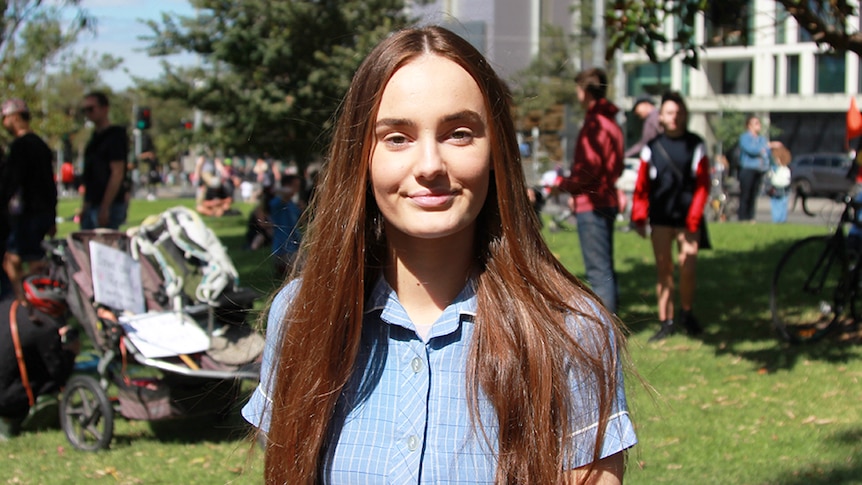 A teenage girl in a school uniform stands in a crowded and sunny park.