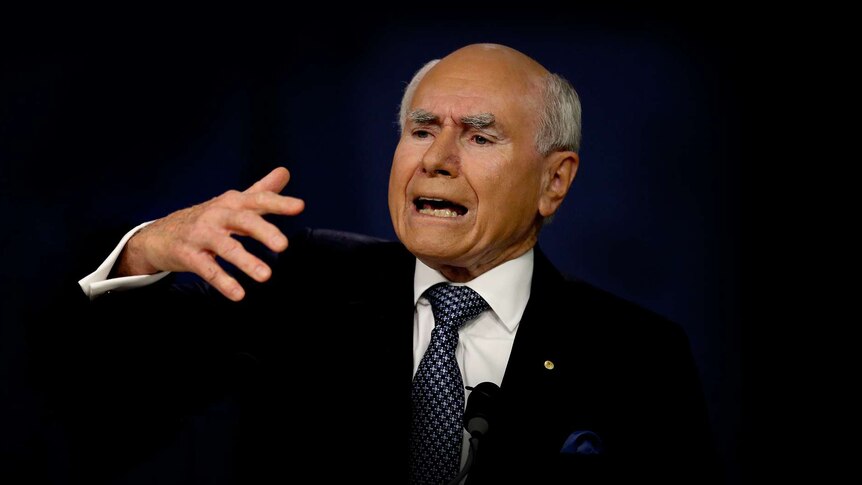 John Howard, eyebrows knitted together, sticks his right hand out while speaking at a microphone. The background is black.
