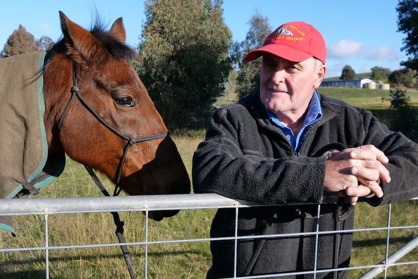 Een oudere man met een rode pet staat bij een boerderijpoort met een bruin paard.