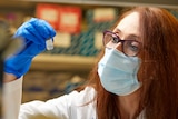 A woman with red hair and black rimmed glasses peers at a vial. She is wearing a surgical mask and gloves