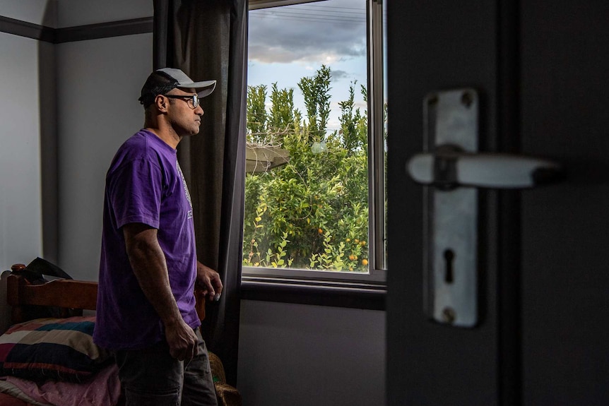 Alifarete Tuitakali in seen through a half-open door, standing near his bed and looking out a window.