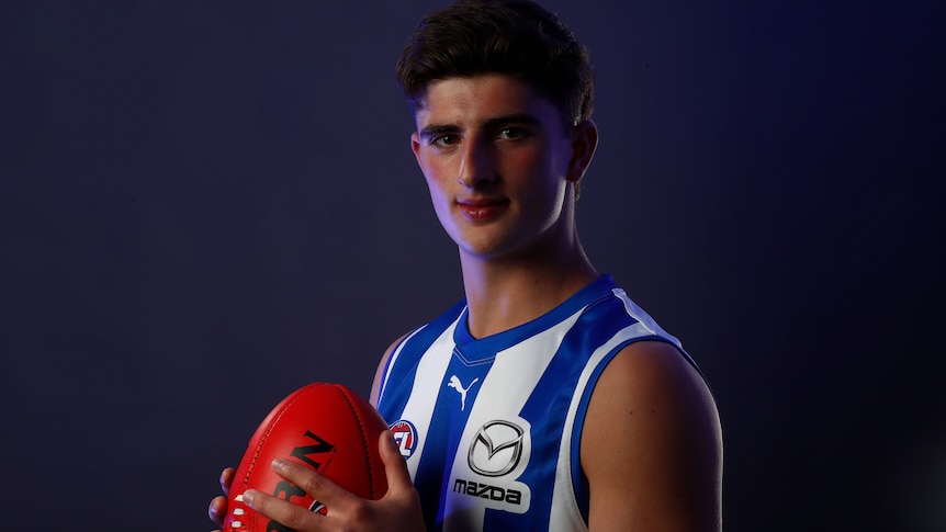 Harry Sheezel poses for a photo while holding a football and wearing a North Melbourne jersey