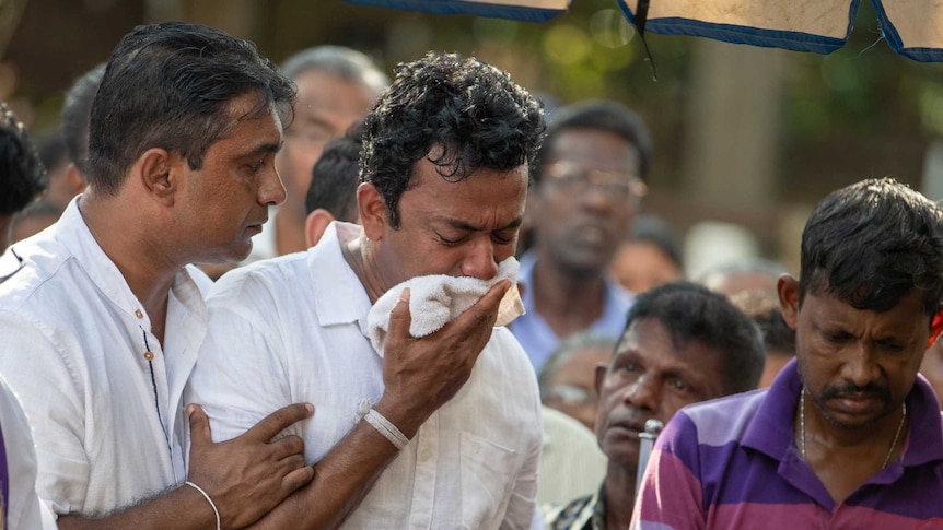 Sudesh Kolonne holds a white cloth to his face and cries surrounded and supported by friends as a priest reads.