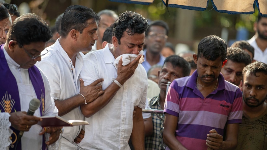 Sudesh Kolonne holds a white cloth to his face and cries surrounded and supported by friends as a priest reads.