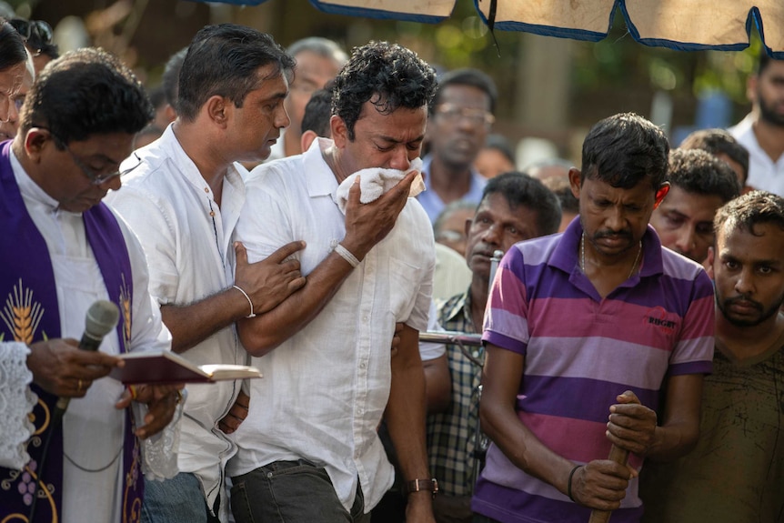 Sudesh Kolonne holds a white cloth to his face and cries surrounded and supported by friends as a priest reads.