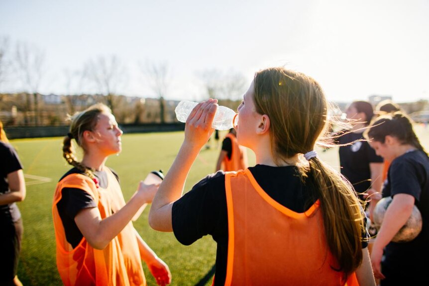 Group of red-faced girls in sports gear on green field drink from water bottles. One holds a soccer ball.