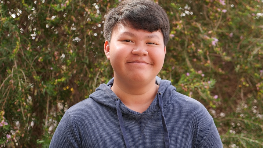 Aldriech looks to the camera and smiles while standing in front of a shrub. He has short dark hair and wears a navy hoodie.