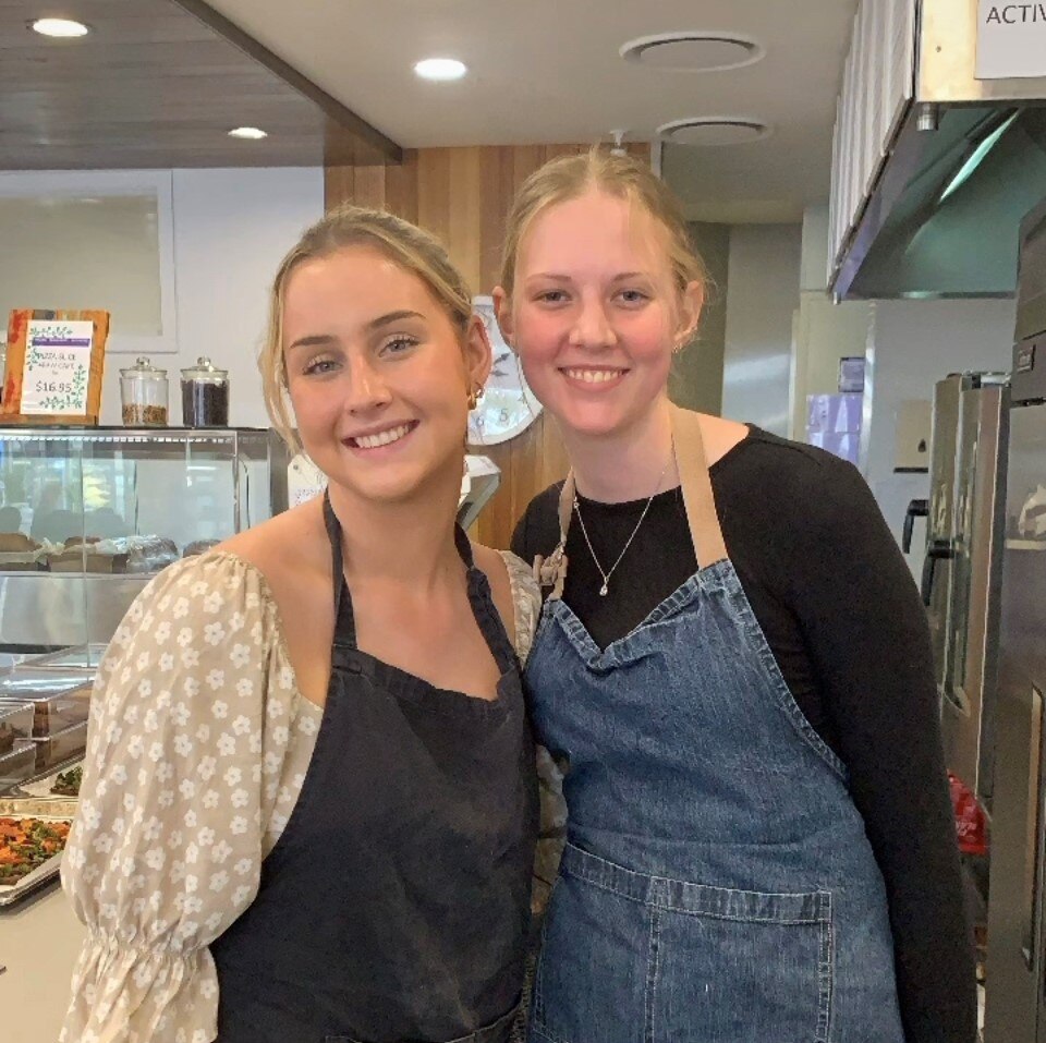 Two blonde women smile at the camera, wearing aprons in a cafe