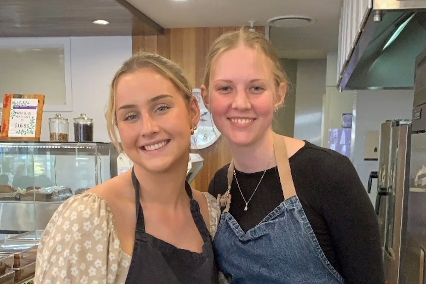 Two blonde women smile at the camera, wearing aprons in a cafe