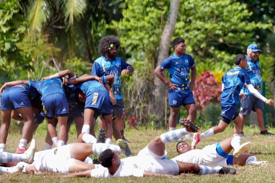 A group of rugby union players in blue uniforms stand and do training moves while a team in white lay on the grass stretching