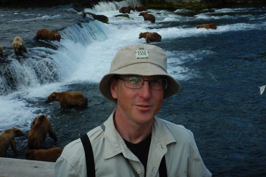 A man in a bucket hat with glasses smiles, as multipe brown bears feed on salmon in the river behind