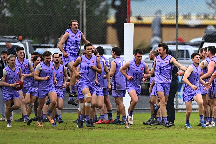 A group of footballers wearing purple jumpers running on to an oval. 