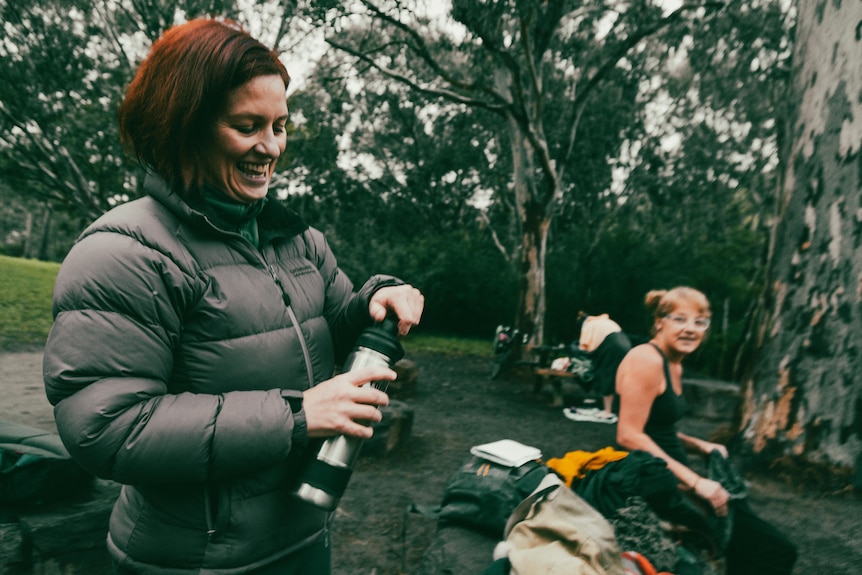 A woman with red hair opens a drink bottle. She is wearing a puffer jacket and smiling.