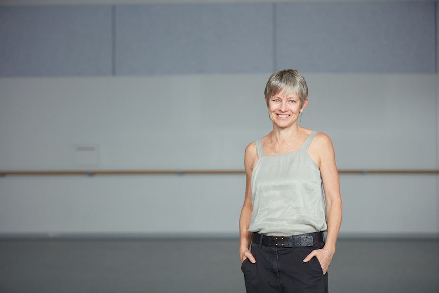 An older fit woman with short silver hair stands in a ballet studio smiling.