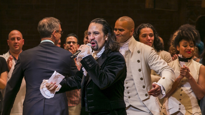 Lin-Man Manuel as Hamilton, holding a microphone, addresses the crowd on the opening night