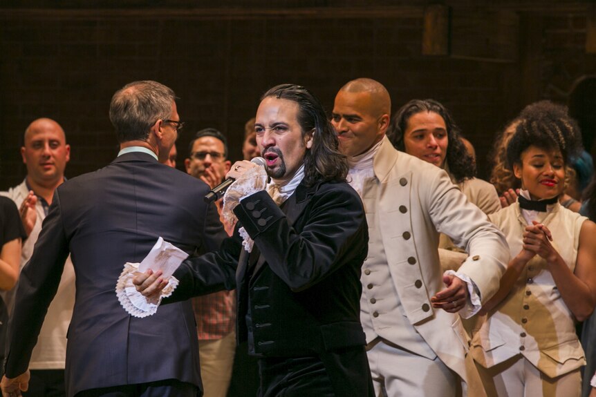 Lin-Manuel Miranda as Hamilton holding a microphone to address the crowd at opening night