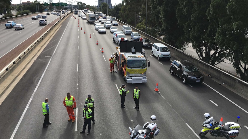 A line of vehicles backed up on the Mitchell Freeway with workers in hi vis gear in the foreground.